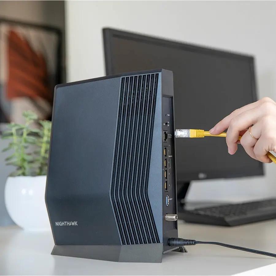 NETGEAR Nighthawk CAX80 black WiFi 6 router with vertical fins on desk; hand plugs Ethernet cable