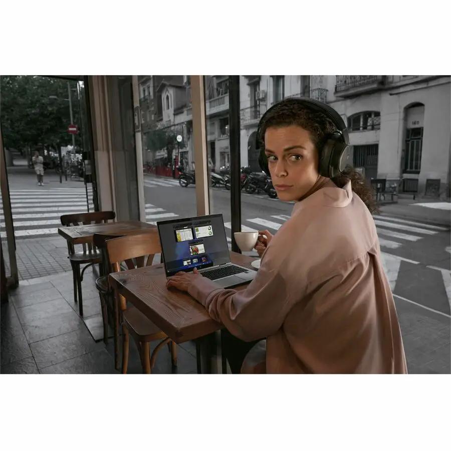 HP EliteBook notebook (silver) on a wooden cafe table by a window with city view; person wearing headphones using laptop.