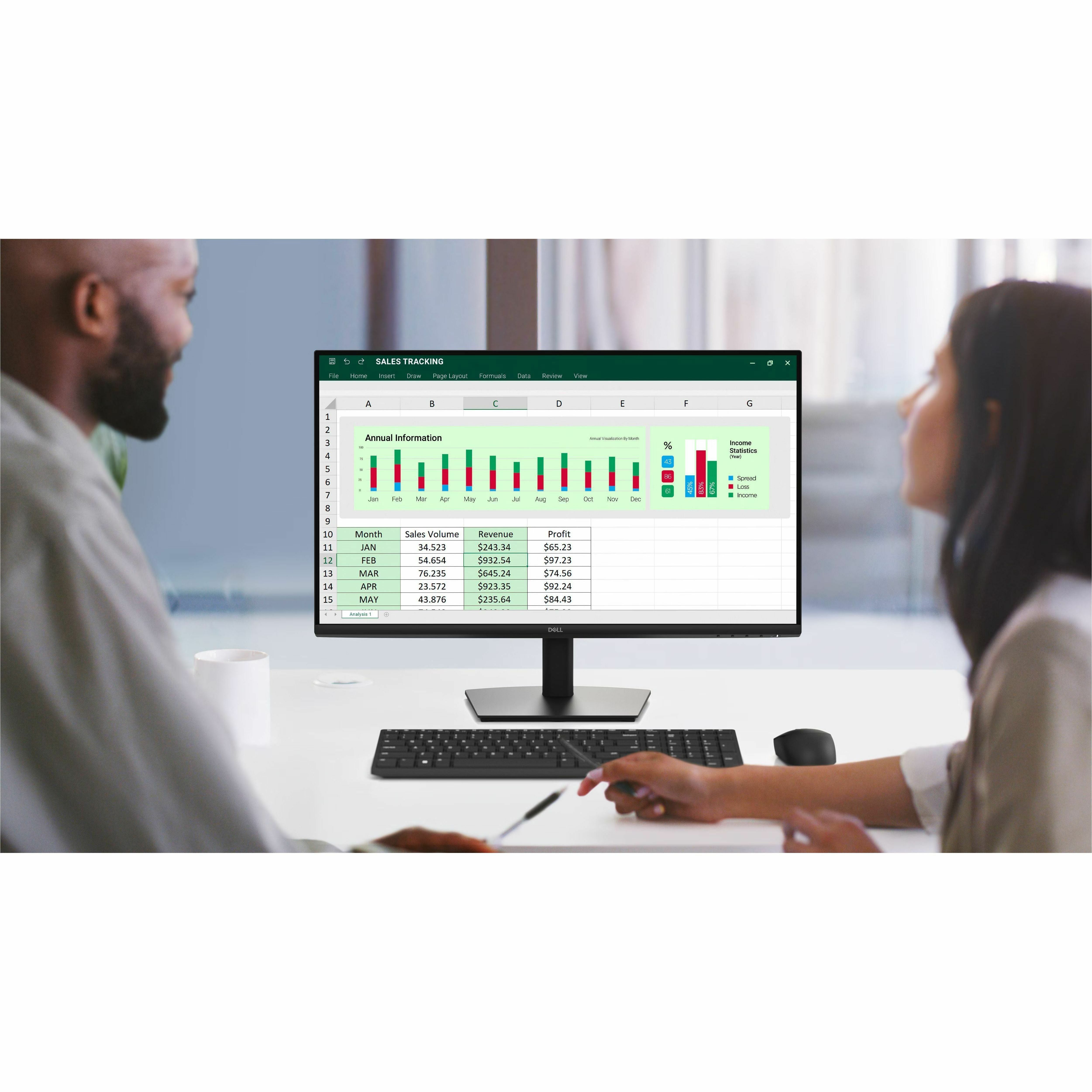 Two people working at a desk with a computer displaying a spreadsheet with charts and data.