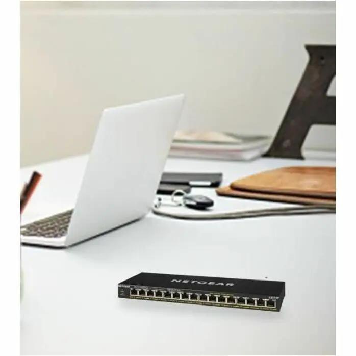 NETGEAR GS316P 16-port Gigabit PoE unmanaged switch, black, on a white desk beside a laptop and notebook.