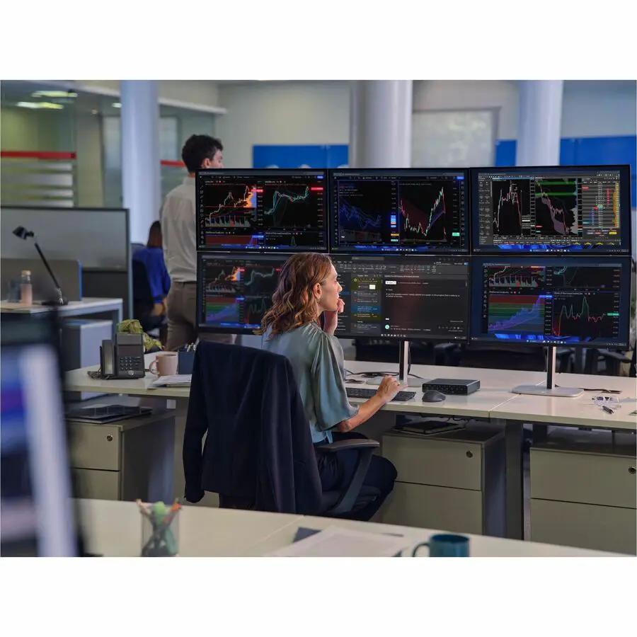 Desktop workstation with six monitors displaying stock charts; a woman sits at a desk in an open-plan office.